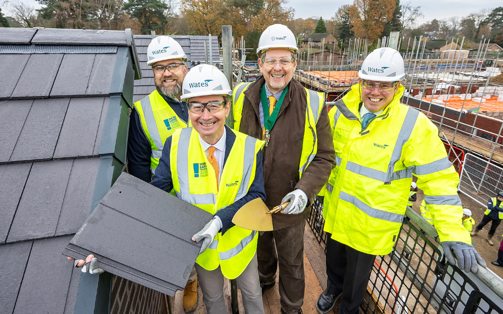 four people in high vis jackets and hard hats smile for the camera while holding up roofing slates on the top of a newly built house