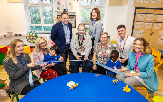 A group of adults smile and cut a ceremonial ribbon while sitting at a children's table inside the new school