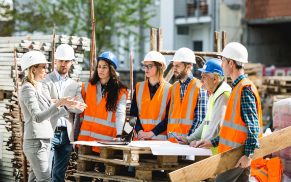 A group of seven construction workers wearing high-viz jackets and hard hats stand round a pile of pallets talking