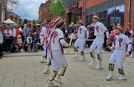 A group of Morris dancers perform on a paved area at Elms Field, Wokingham, on a sunny day in front of a large cheering crowd