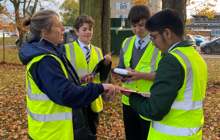 A group of children and one adult in high vis jackets gather around and look at air quality measuring devices they're holding