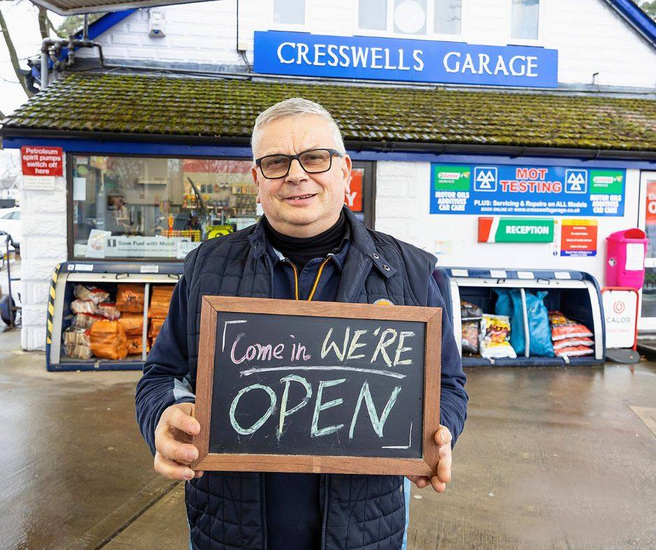 The owner of Cresswells Garage standing outside his business with a sign saying "we're open"