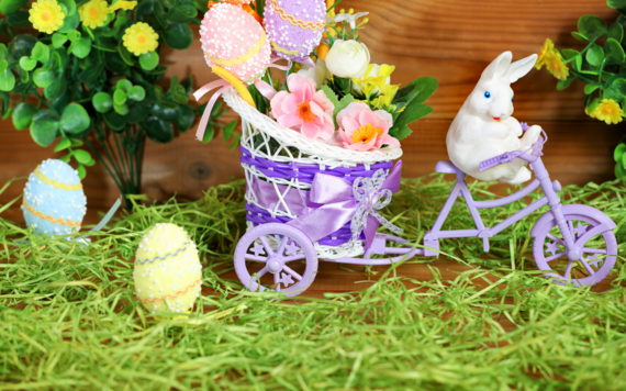 Toy bunny on a bike with a basket filled with easter eggs. In a garden surrounded by greenery and more easter eggs on the grass.