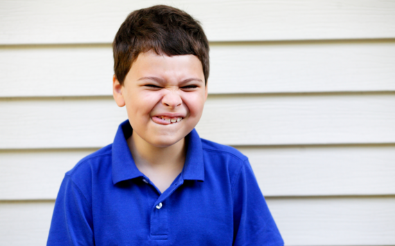 A young boy wearing a dark blue polo shirt standing against a white wall screws up his face in excitement and pokes his tongue out