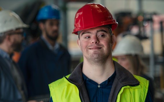 A young man with down syndrome wearing a hard hat and high viz jacket on a bulding site