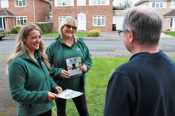 Two women in dark green council jackets chat with a man on his doorstep while holding information leaflets about recycling
