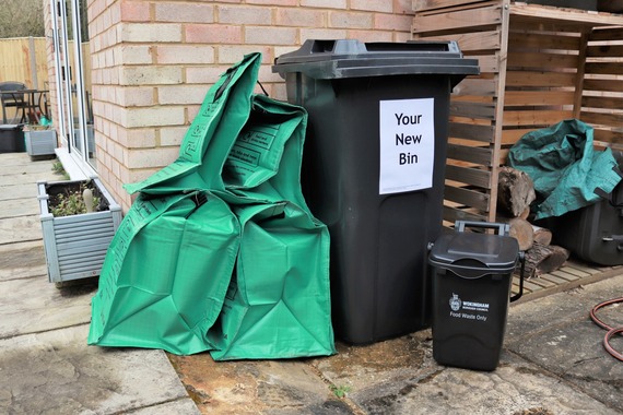 Four green recycling bags, a black recycling bin with a sign saying "your new bin" and a food waste bin behind a house