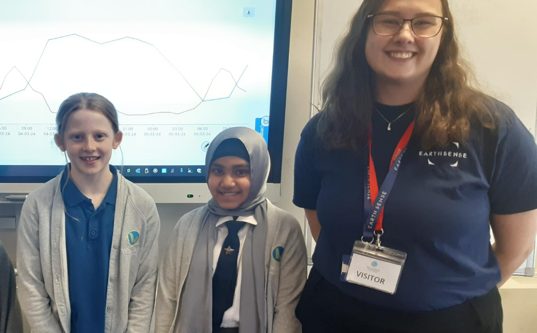 two schoolgirls and a young woman smile in front of a board displaying an air pollution data graph