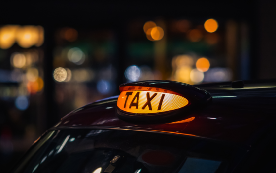 The roof of a black taxi with a yellow taxi light lit up against a blurred backdrop of lights