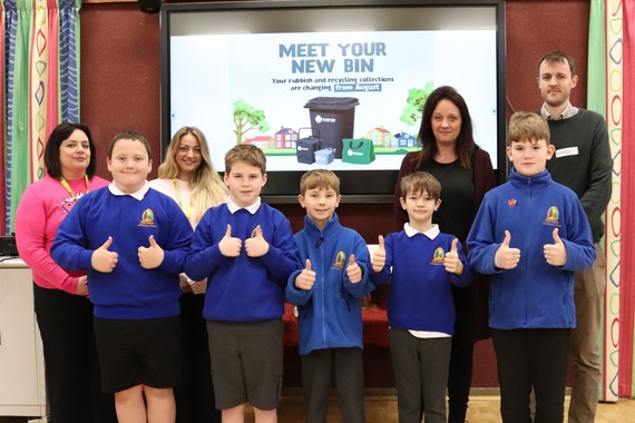 Our recycling officers with a group of children smiling and making thumbs-up to the camera, with a "meet your new bin" presentation behind them