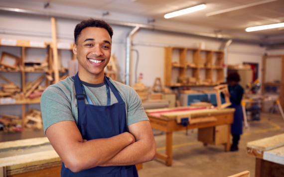 A young man smiling in an apron, against a background of woodworking tools