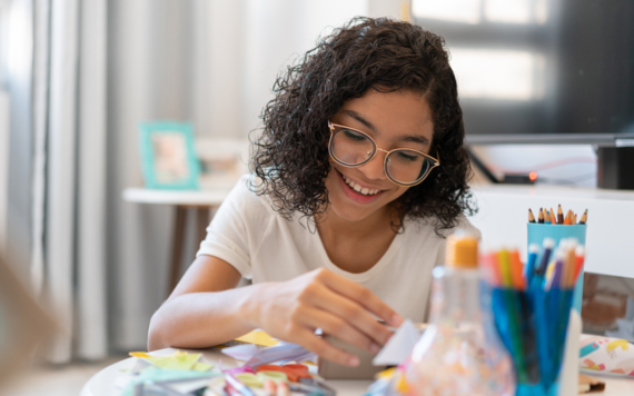 A girl with dark curly hair and glasses, smiling at a table with art supplies and colouring pencils 