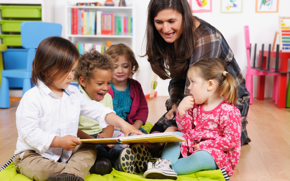 A nursery teacher with four young children crowded around a book