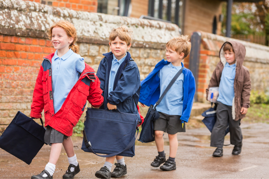Four young children wearing schools uniforms and coats and carrying book bags walk along a pavement