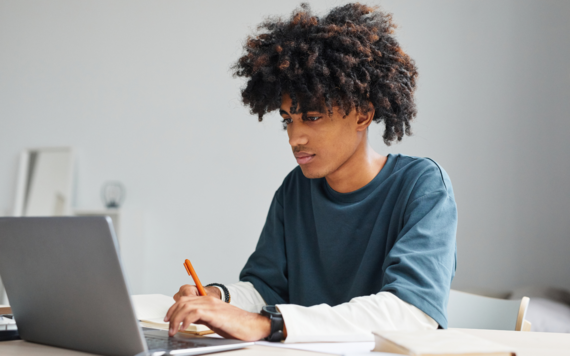 A teenage boy sitting at the table with a laptop and pen