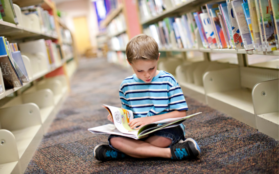 A boy sitting crossed legged on the floor in the library, reading a picture book