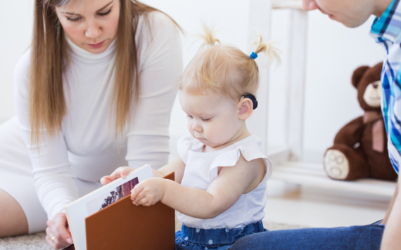 A young toddler wearing hearing aids looking at a book with two adults