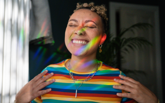 Image of a woman in a rainbow jumper smiling with light shining through the window on her