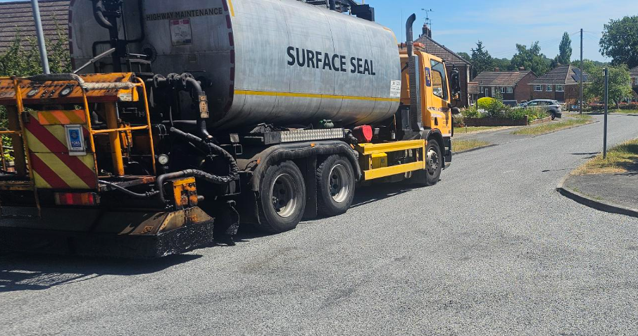 A large lorry marked "SURFACE SEAL" drives along a road spraying a substance from a trailer at the back