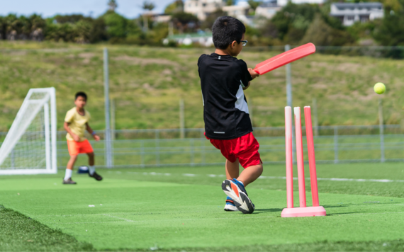 A boy swinging a red cricket bat to hit a ball