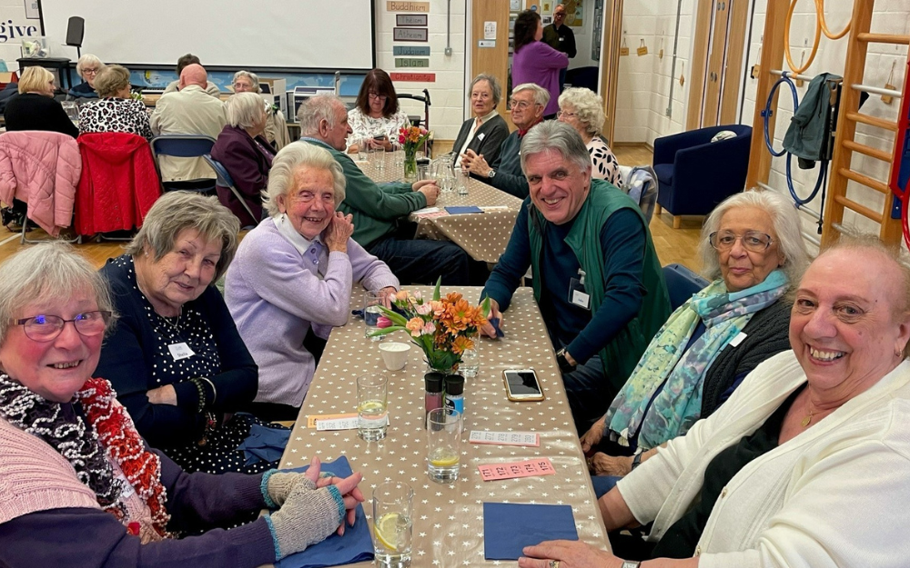 A volunteer friendship table run by Link Visiting Scheme, six older people smile to camera sat around a table
