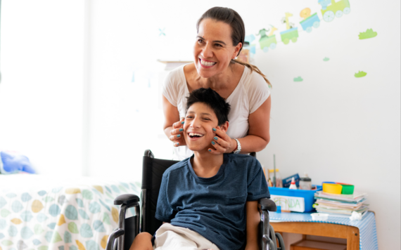 A boy in a wheelchair laughing while his mum stands behind him and rests her hands on his face