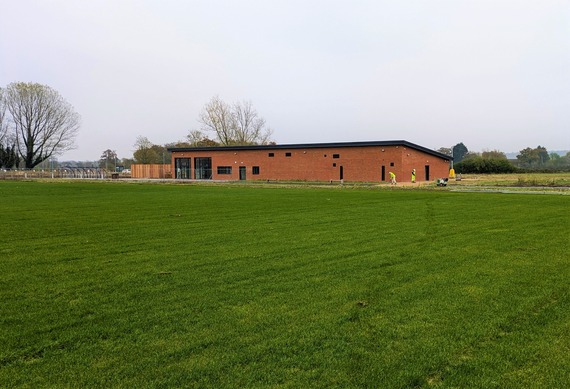 A newly built single storey sports pavilion in the background and a newly planted playing field in the foreground at High Copse