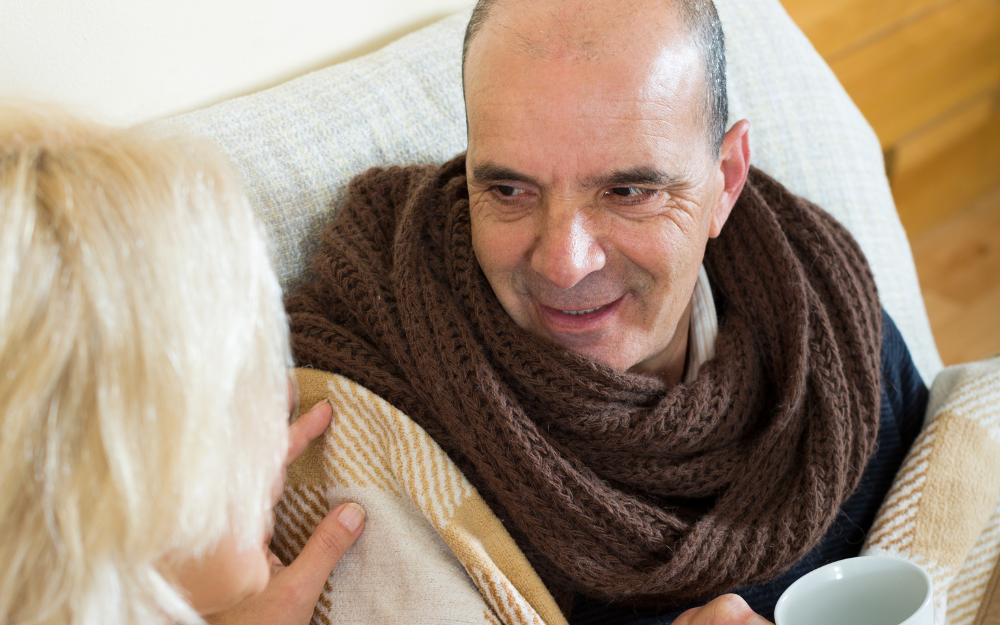 A woman comforts a cold older man wrapped up in scarfs with a warm drink