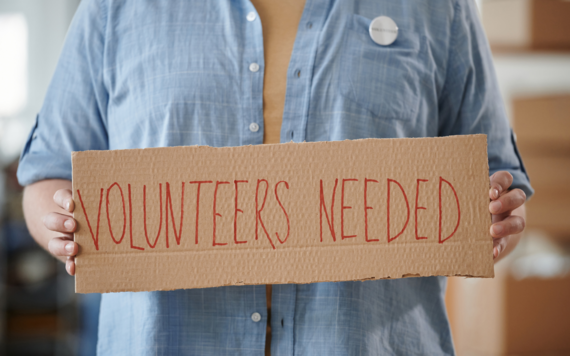 A close up of a sign held by a person. The sign reads Volunteers Needed 