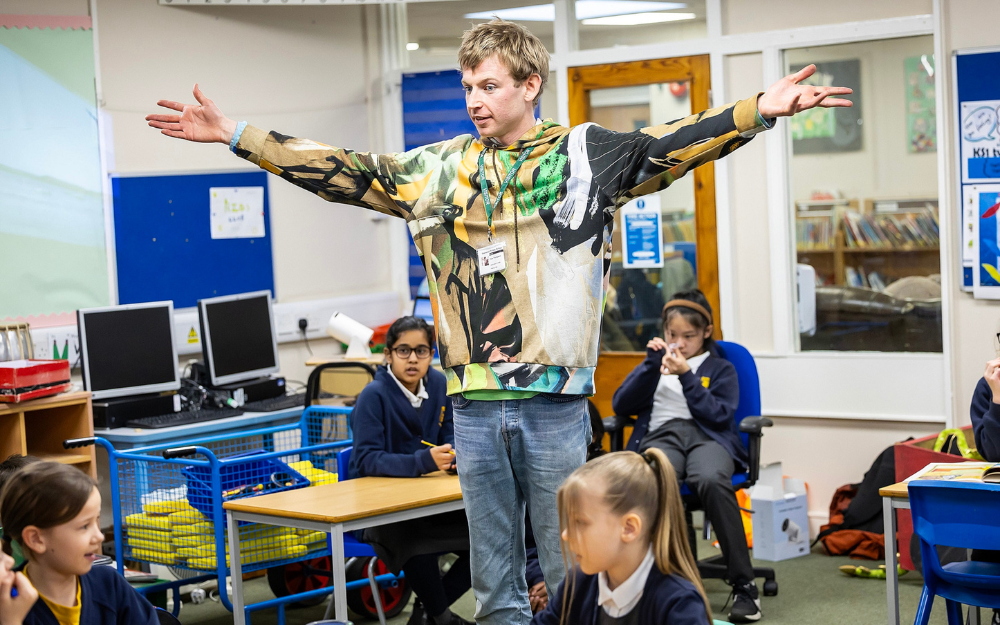 A man stands in a classroom with his arms out, engaging the pupils