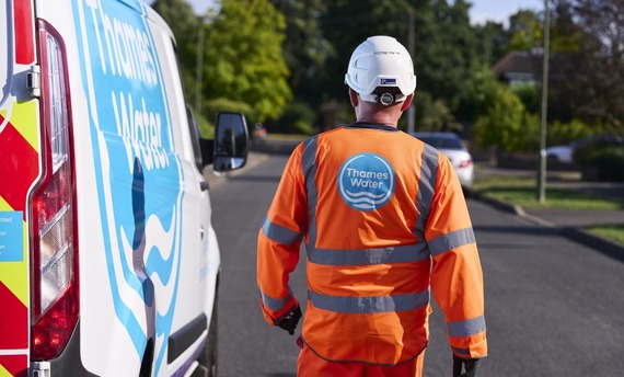 The back of a Thames Water engineer wearing high viz orange overalls with the company logo, standing in the road next to a branded van