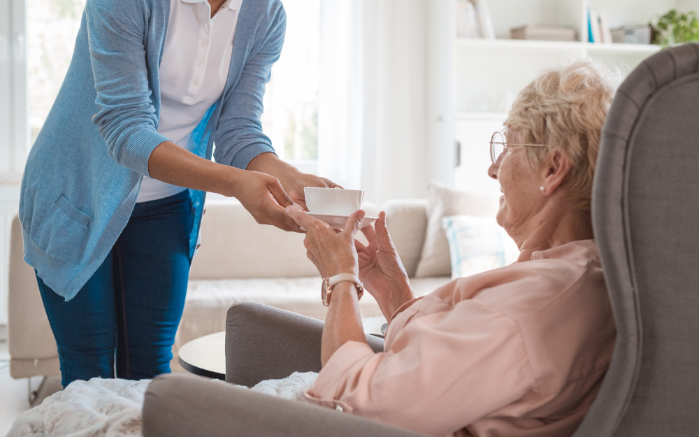 An older woman in a care home receives a cup of tea from a care worker