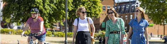 cropped image showing three women walking and smiling with another woman behind them on a bicycle