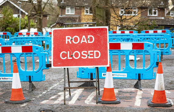 A road closed sign with barriers and a newly treated road behind it