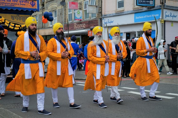a row of men in traditional Sikh attire, including orange robes, yellow turbans and swords, process down a road in East Reading