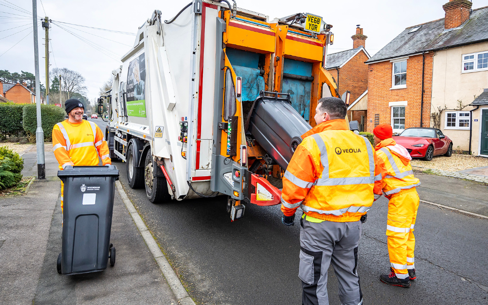 Three waste operatives loading black bins into a waste lorry
