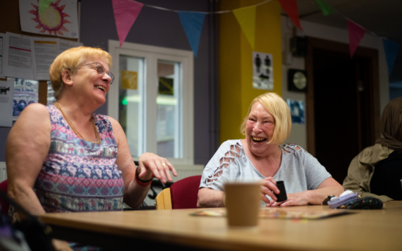 Two ladies laughing together in a community centre with a cup of tea
