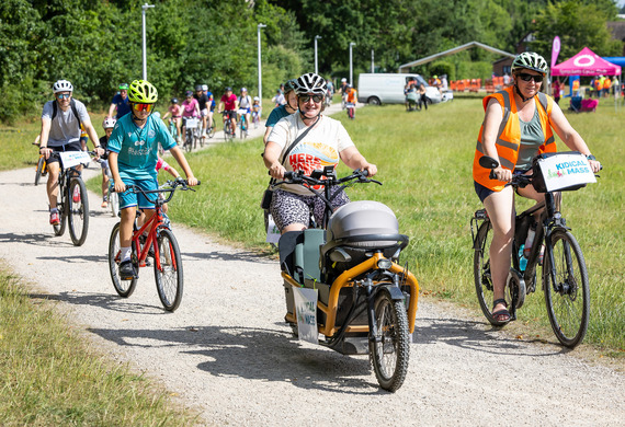 a group of adults and children riding bikes along a narrow path through a field on a bright sunny day
