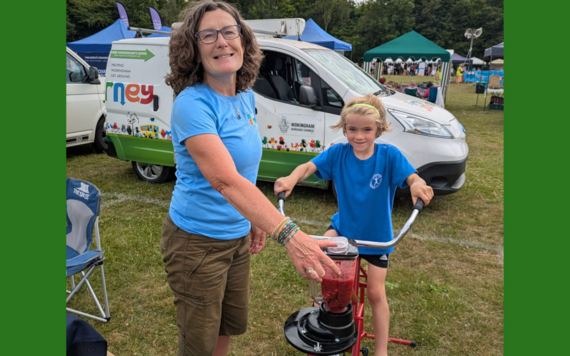 A woman supervises a young girl as she rides a static bike connected to a fruit smoothie maker