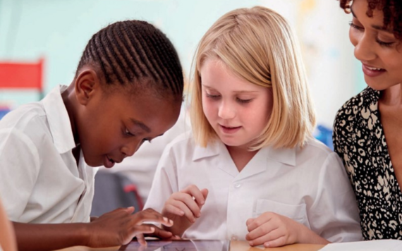 Two children in school uniform using a tablet, sat next to their teacher