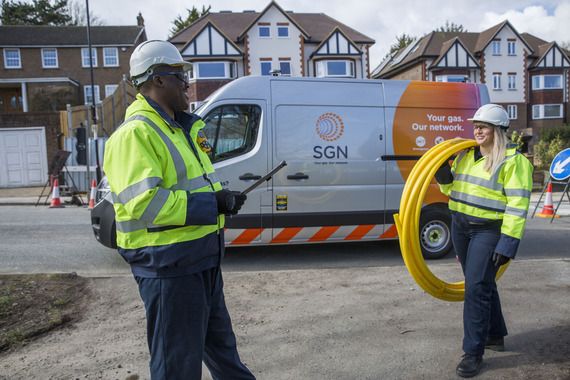 two SGN staff in high vis jackets and hard hats carrying coiled lengths of yellow plastic pipe at a works site in a residential street