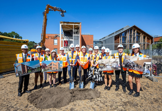 a group of just over a dozen students and adults, all in hard hats and hi vis, posing on a building site holding up drawings of the finished centre