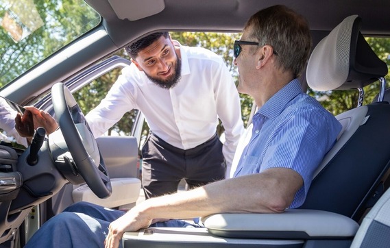 a man sits in the driving seat of an electric vehicle holding the wheel while talking through the window to a young salesman