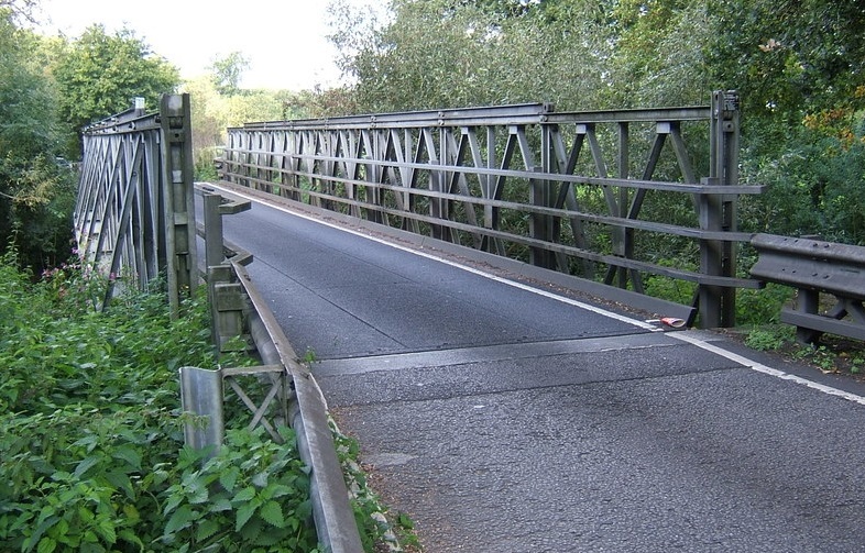 A single-lane metal bridge passing over a small river, with trees and hedges on either side