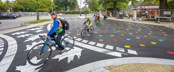 a group of cyclists including children ride across a colourful new junction painted with leaves in different colours
