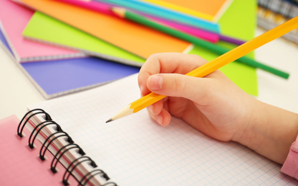 A child's hand holds a pencil above a blank notebook page.