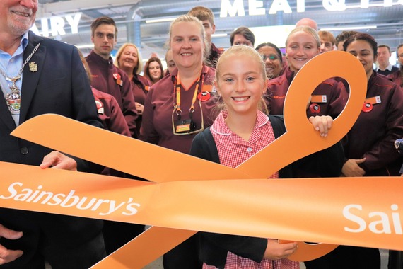 a smiling girl holds a large pair of ceremonial orange scissors up to a branded Sainsbury's ribbon