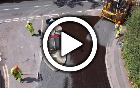 a play button superimposed over an aerial image of workmen in high vis laying a new surface on a road