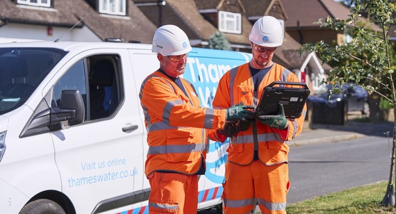 Thames Water engineers in branded high viz and helmets, with a branded van, stand in the road looking at a computer tablet