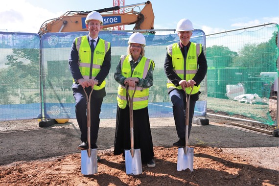 two men and a woman, wearing white hard hats and green hi vis, smile while digging spaces into the ground on a building site